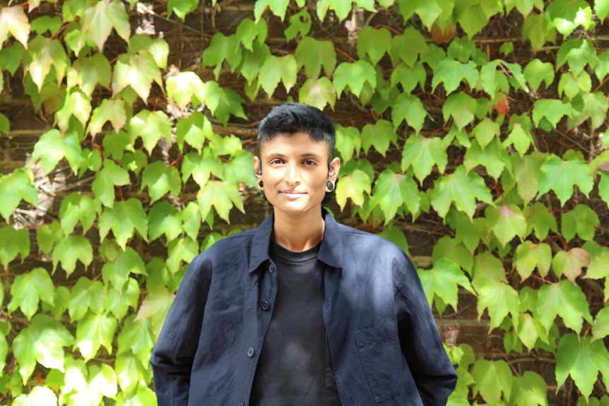 Priya Kunjan - a person with short dark hair and a dark blue shirt standing in front of an ivy-covered wall