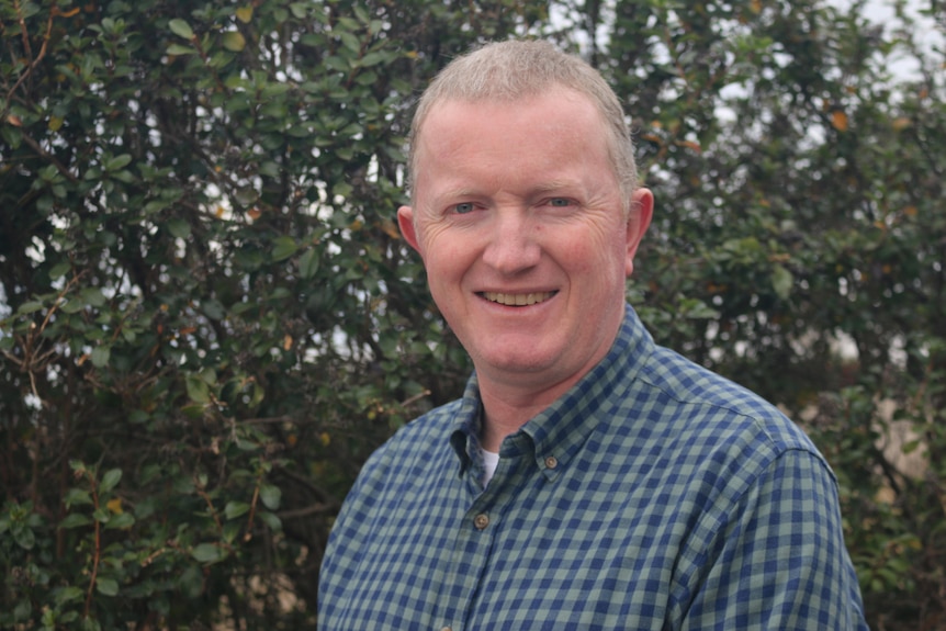 Gene Hodgins - Photo of a middle-aged man in a checkered shirt, smiling, standing in front of a tree.