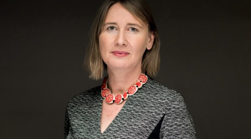 Rebecca Bentley - an older white woman in a gray dress and a gold necklace looks at the camera.